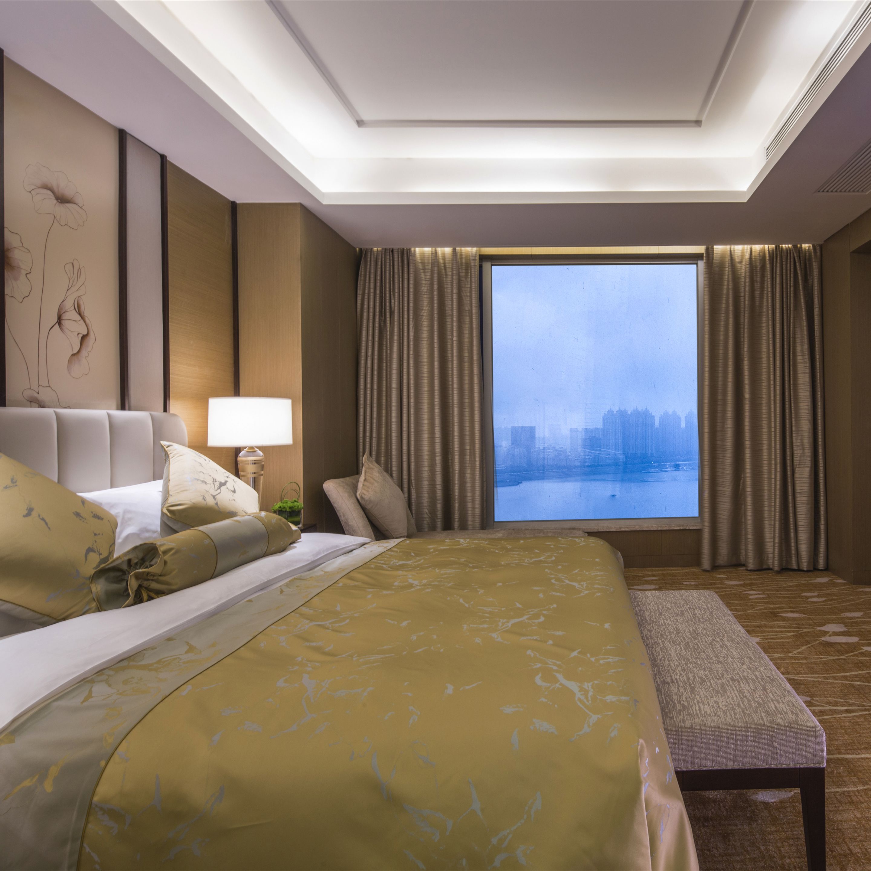 A hotel room with a large bed, a bench, a nightstand, a lamp, and a view of a cityscape through a large window.
