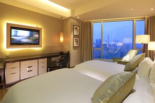 A hotel room with a large bed, a television mounted on the wall, a desk with a chair, and a view of the cityscape through the window.