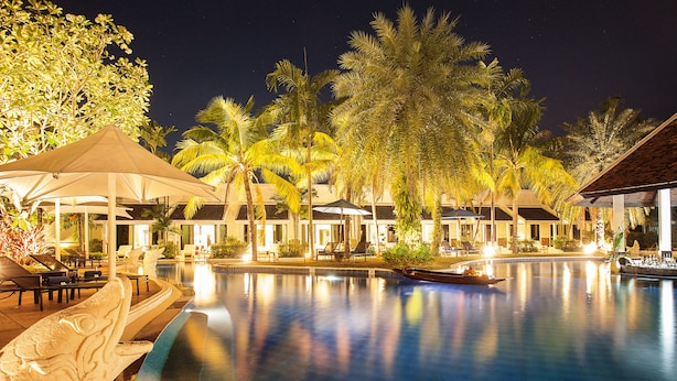 Vista nocturna de un área de piscina en un resort, con palmeras, sillones de descanso y sombrillas.