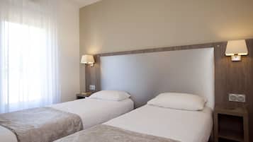 A hotel room with two single beds, a wooden headboard, bedside lamps, a nightstand, and a window with sheer curtains.