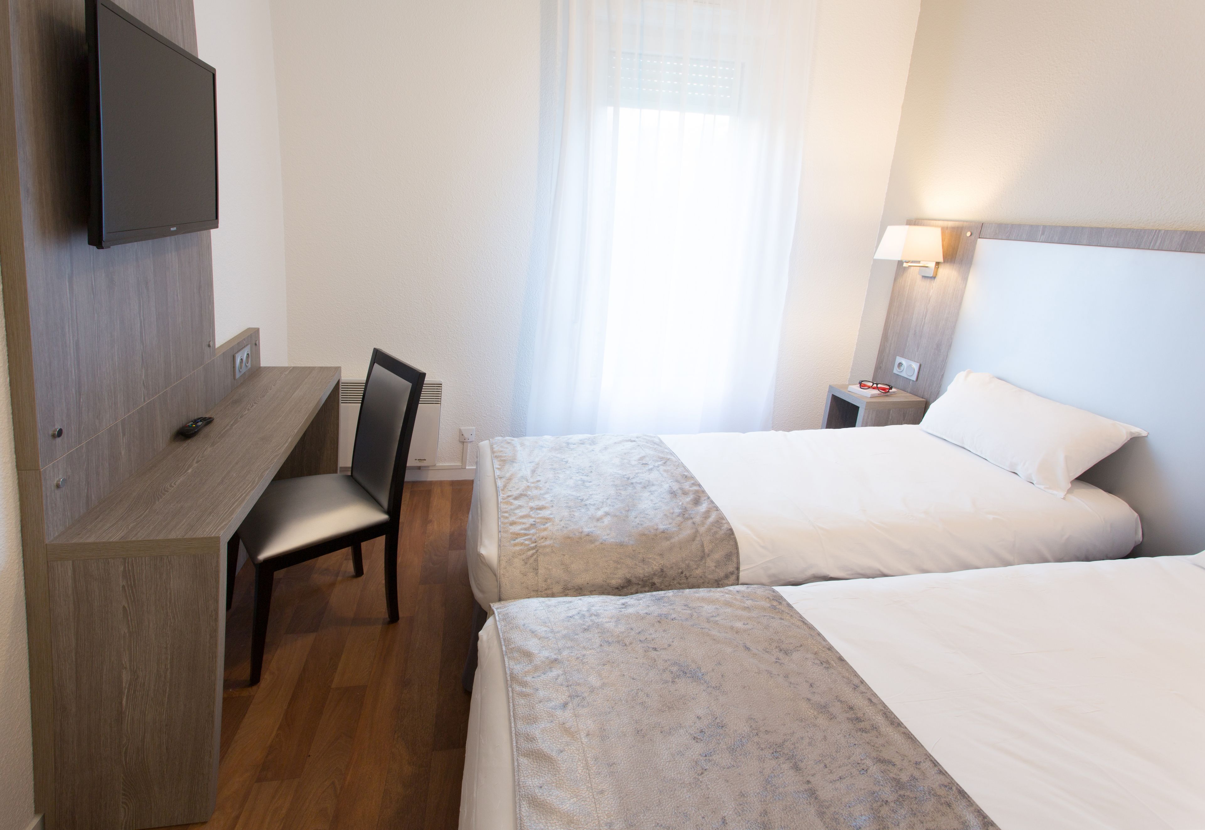 A hotel room with a desk, two beds, a chair, a TV, and a window with curtains.