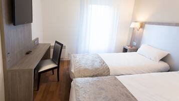 A hotel room with a desk, two beds, a chair, a TV, and a window with curtains.