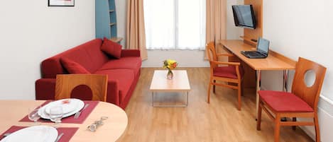 Une pièce meublée avec un canapé rouge, une table à manger dressée avec des assiettes et des couverts, une petite table basse avec un vase, un bureau avec un ordinateur et une télévision, et une fenêtre avec des rideaux.