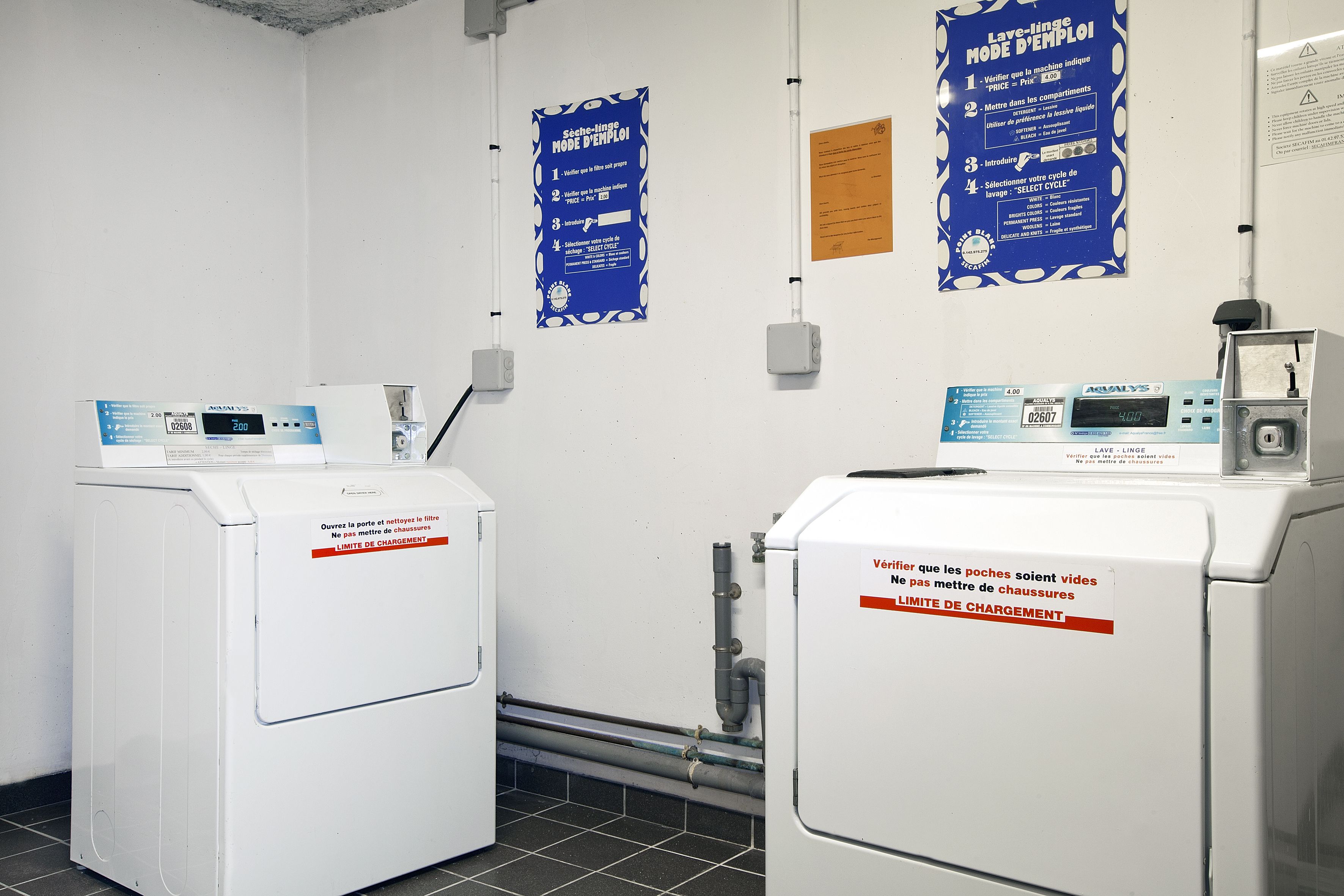 A laundry facility with two washing machines and a dryer, each with a digital display and a control panel. There are instructional posters on the wall and a notice about the washing machines' capacity.