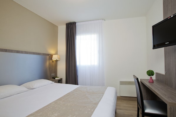 Une chambre d’hôtel avec un lit, un bureau, une chaise, une télévision et une fenêtre avec des rideaux.