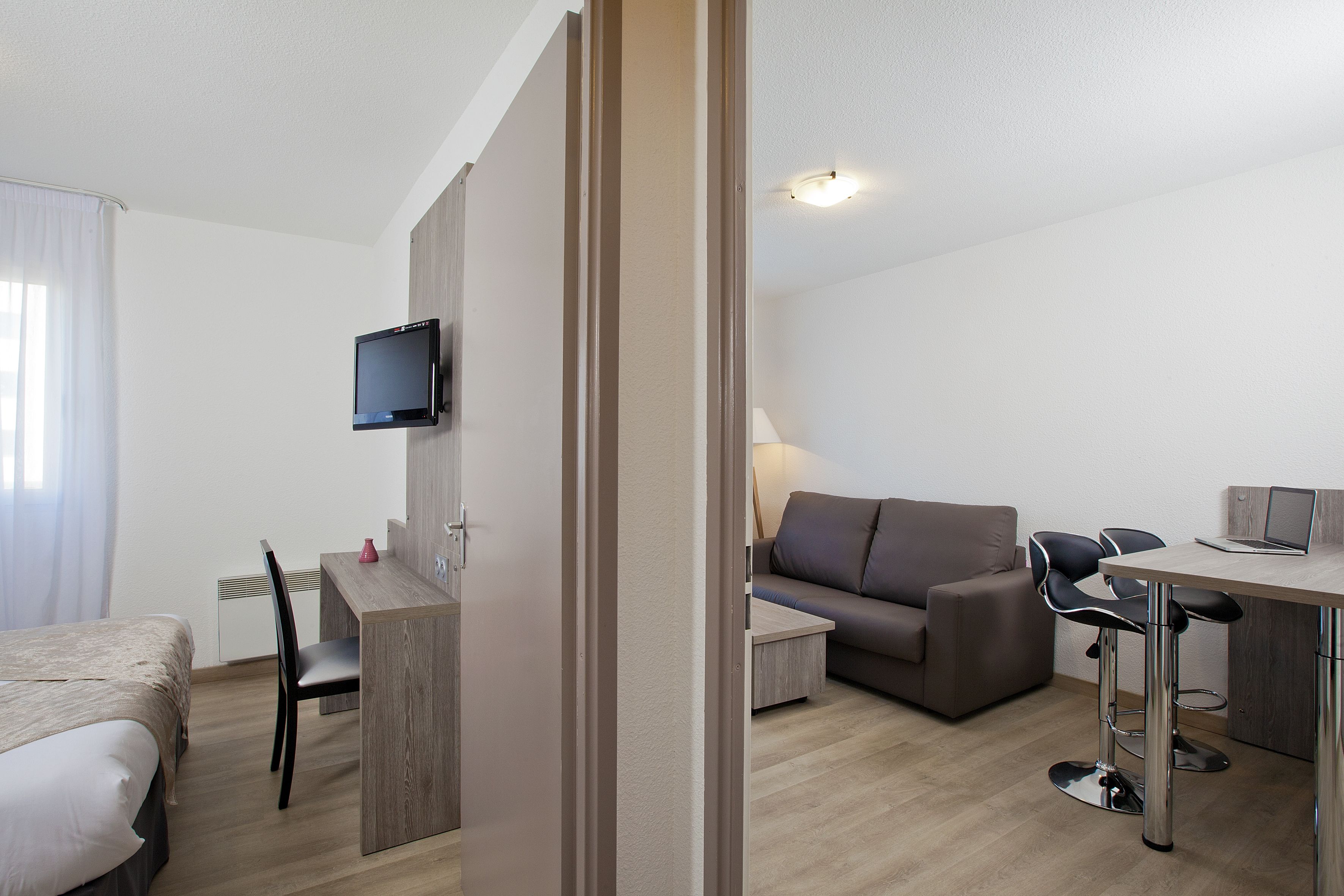 A modern hotel room with a bed, a desk, a TV mounted on the wall, a sofa, and a dining area with chairs.