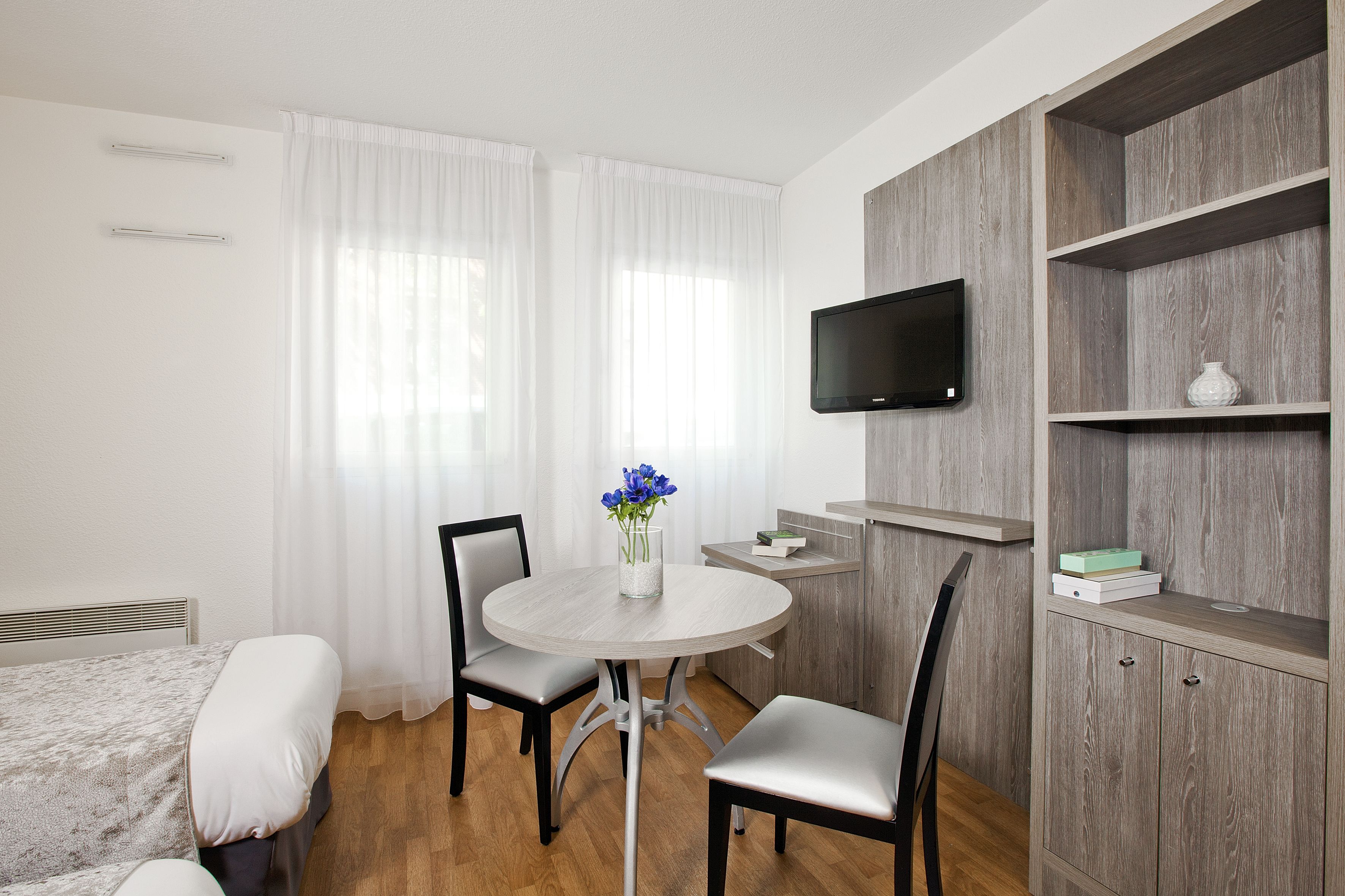 A hotel room with a bed, a round dining table with chairs, a TV mounted on the wall, and a wooden wardrobe.