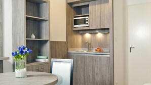 Een moderne keuken met houten kastjes, een magnetron en een spoelbak. Er is een eethoek met een ronde tafel en stoelen, en een bed in de hoek.