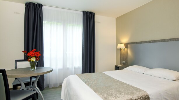 Een hotelkamer met een bed, een tafel met een bloemenvaas, een stoel en een raam met gordijnen.