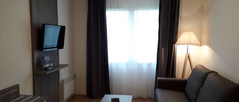 Habitación de hotel con televisión de pantalla plana, sofá, mesa de centro, lámpara y ventana con cortinas.