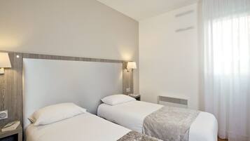 Habitación de hotel con dos camas, una mesita de noche con lámpara, un calentador en la pared y una ventana con cortinas.