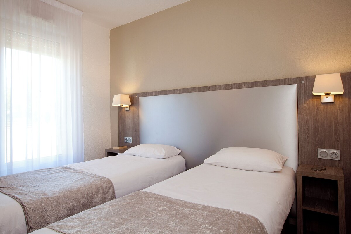 Habitación de hotel con dos camas individuales, cabecera de madera, lámparas de noche, mesitas de noche y una ventana con cortinas traslúcidas.