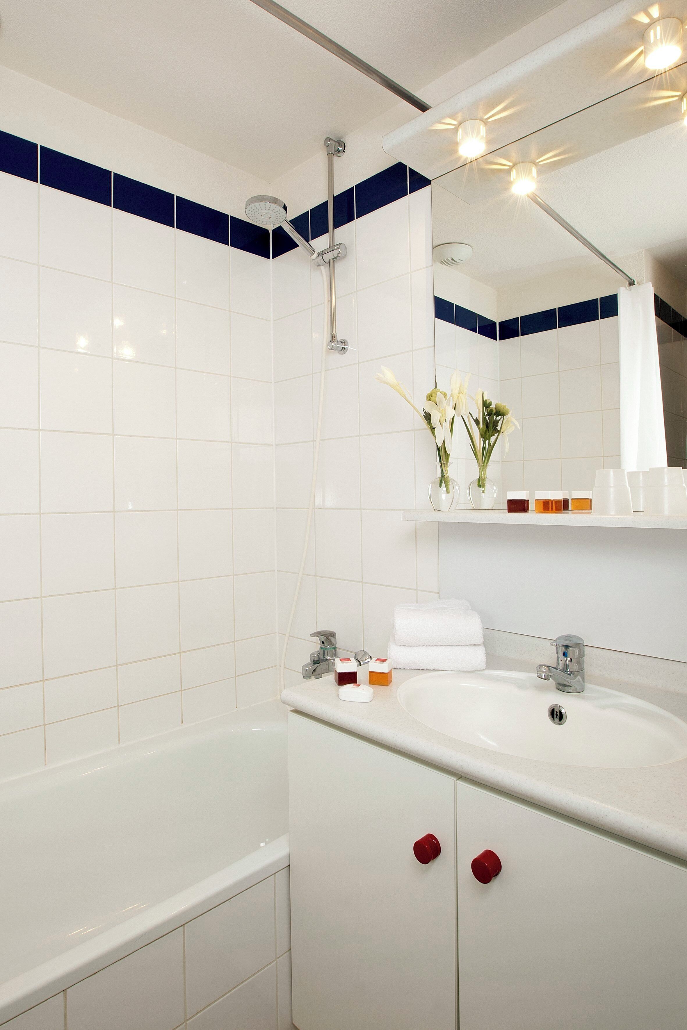 A bathroom with white tiles, a bathtub, a sink with a mirror above it, and a shelf with toiletries.
