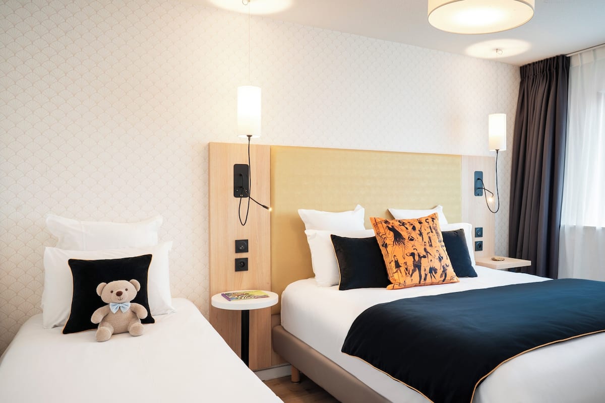 A hotel room with two beds, a nightstand with a lamp, a teddy bear on one bed, and a window with curtains.