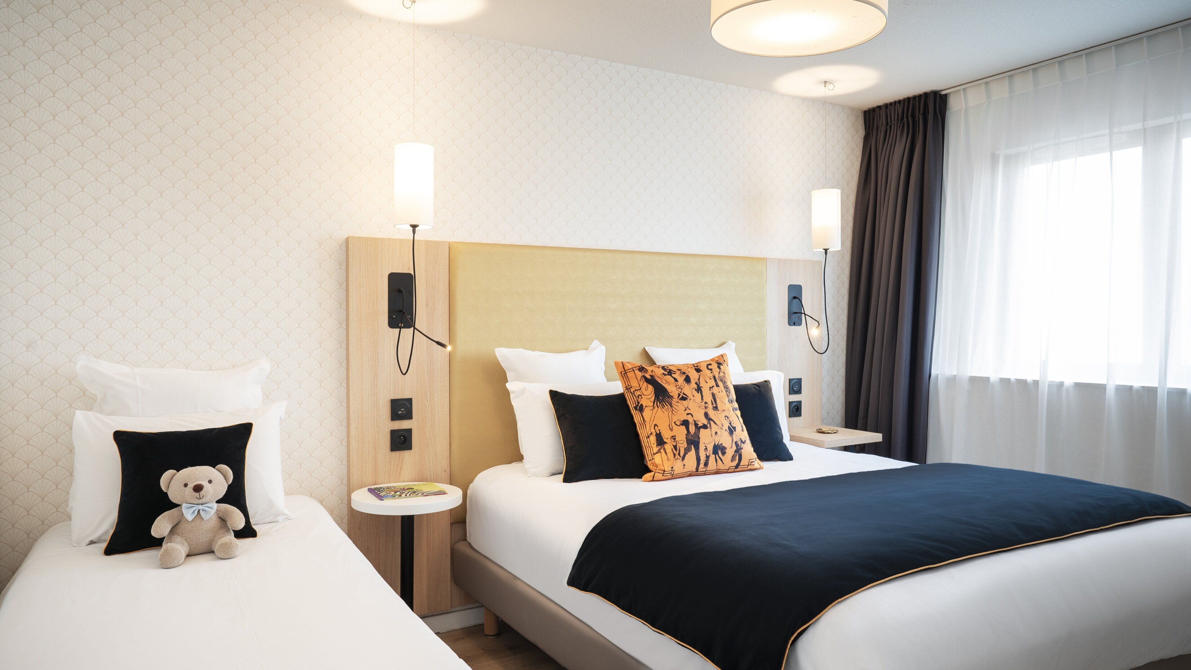 Une chambre d’hôtel avec un grand lit, deux tables de chevet, un ours en peluche sur l’un des lits et une lampe fixée au mur.