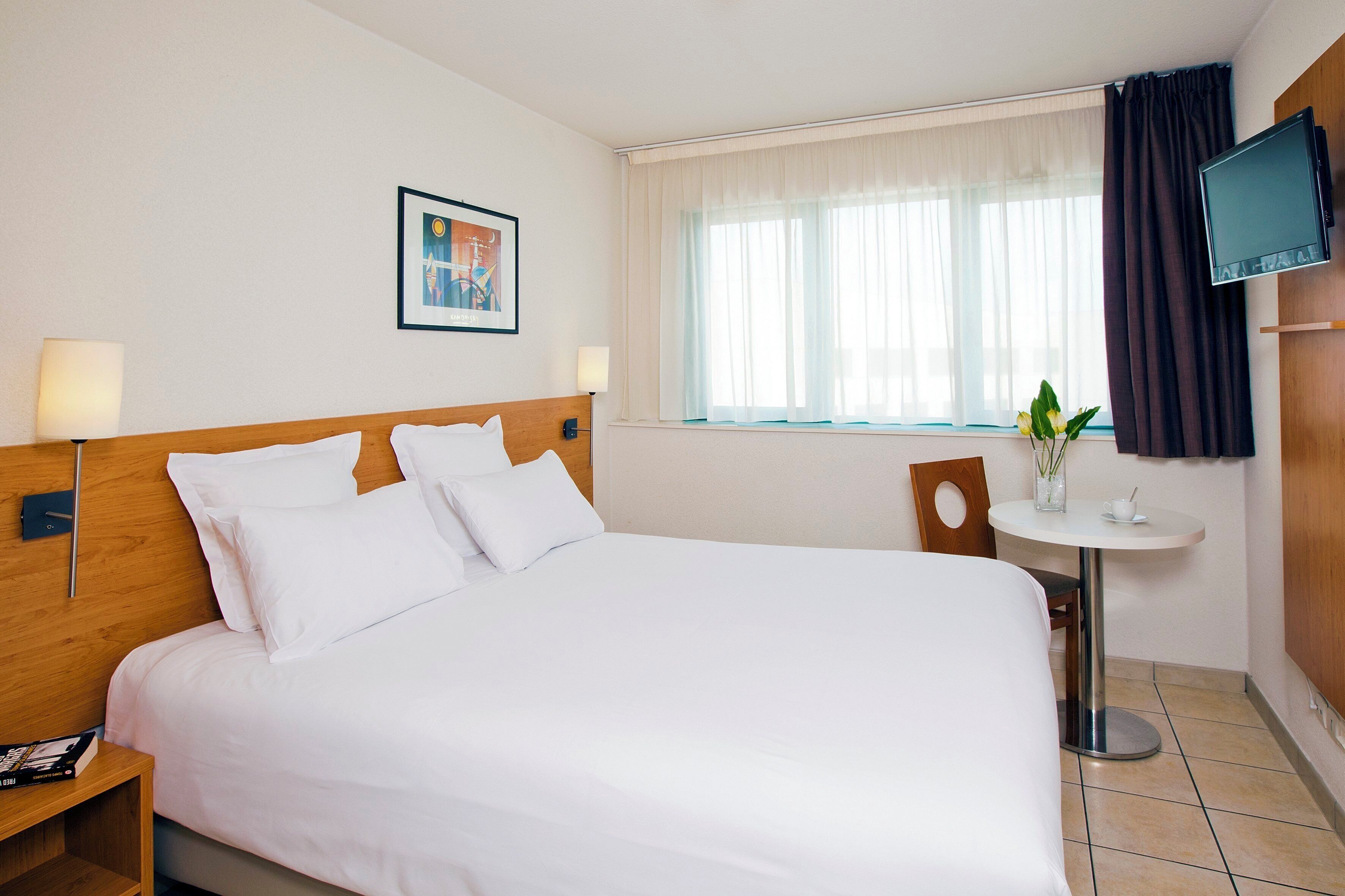 Une chambre d’hôtel avec un grand lit, une table de chevet, une petite table ronde ornée d’une plante, un téléviseur fixé au mur et une fenêtre avec des rideaux.