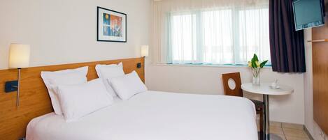 Una habitación de hotel con una cama grande, una mesita de noche, una pequeña mesa redonda con una planta, un televisor montado en la pared y una ventana con cortinas.
