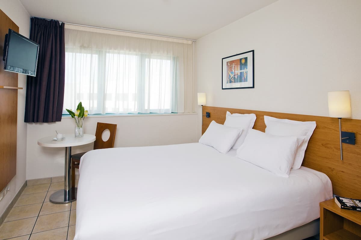 A hotel room with a large bed, a small round table, a TV mounted on the wall, and a window with curtains.