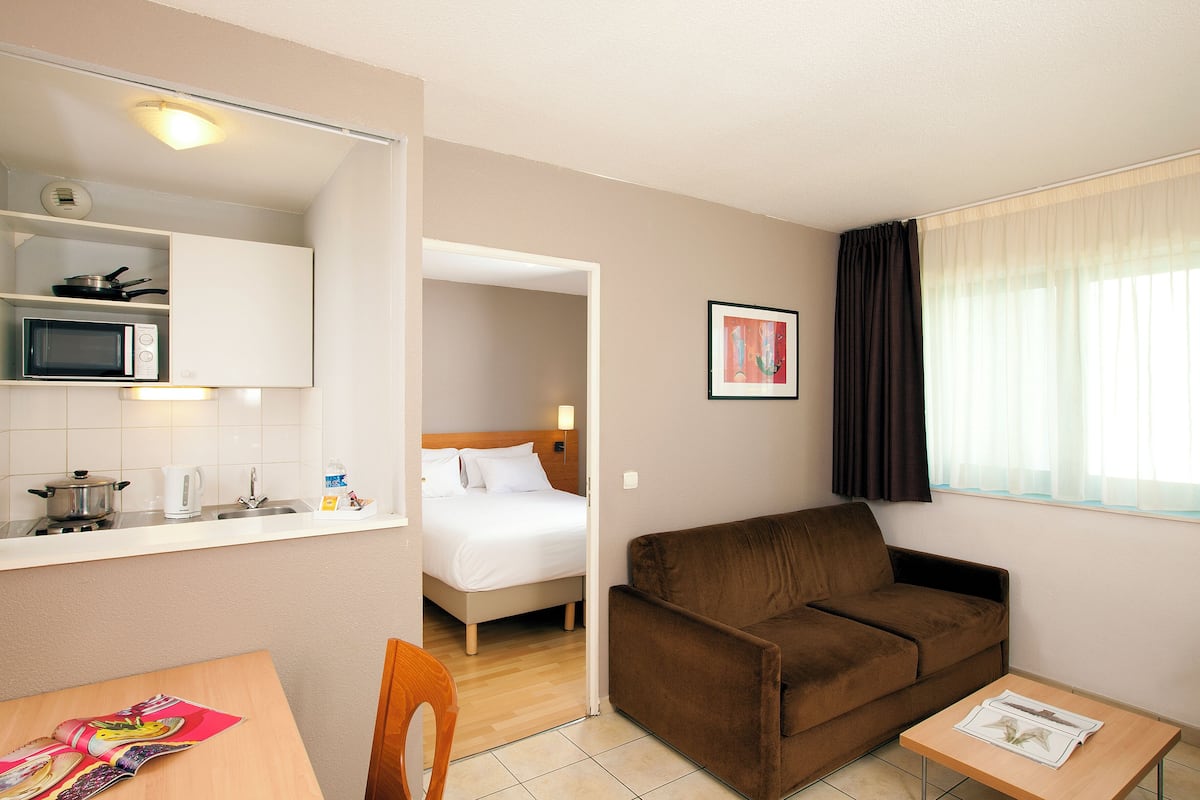A compact hotel room with a kitchenette, a bed, a brown sofa, a small table, and a window with curtains.