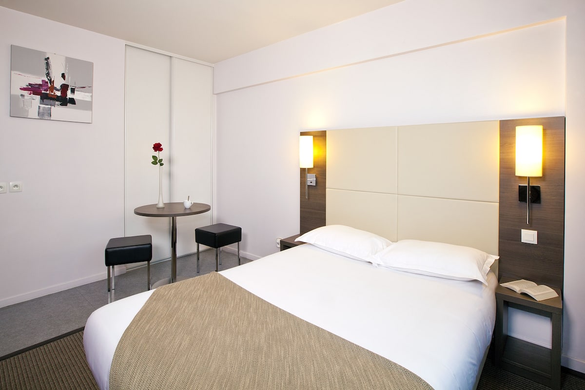 A hotel room with a large bed, bedside tables, a small round table, and a chair.