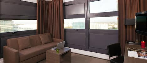 Una habitación de hotel moderna con un sofá marrón, una mesita de madera con un ordenador portátil, una silla negra y un televisor.