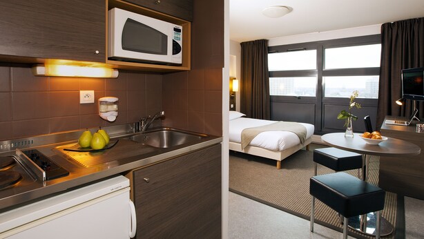 A hotel room with a kitchenette, a bed, a dining area, and a view of the city.
