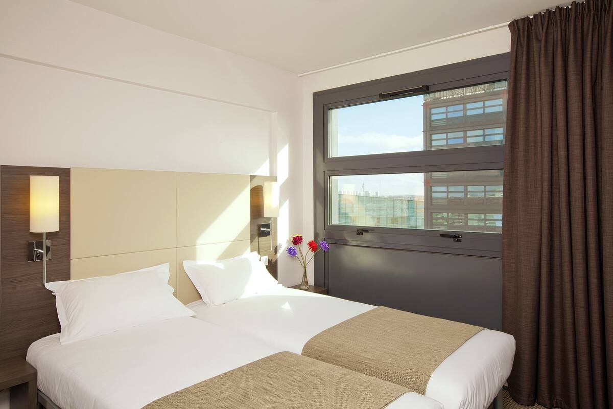 A hotel room with a large bed, a nightstand, a lamp, a window with curtains, and a vase with flowers.