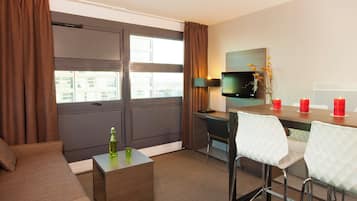 Habitación de hotel con sofá, una pequeña mesa con una botella, escritorio con computadora, televisor y zona de comedor con sillas y velas.