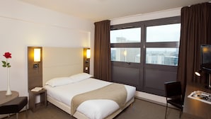 A hotel room with a bed, bedside table, lamp, and a view of the city through a large window.