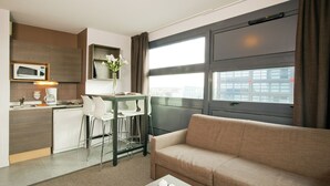 A modern hotel room with a kitchenette, a sofa, and a large window.