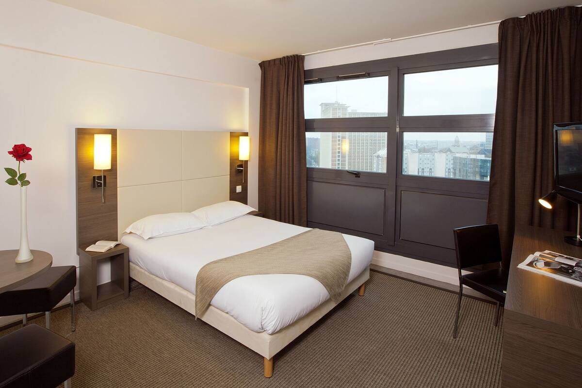 A hotel room with a bed, bedside tables, a desk, a chair, a TV, and a window with curtains.