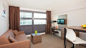 A hotel room with a brown sofa, a small wooden side table, a white chair, a desk with a computer, a flat-screen TV, and a bowl of fruit on the desk.