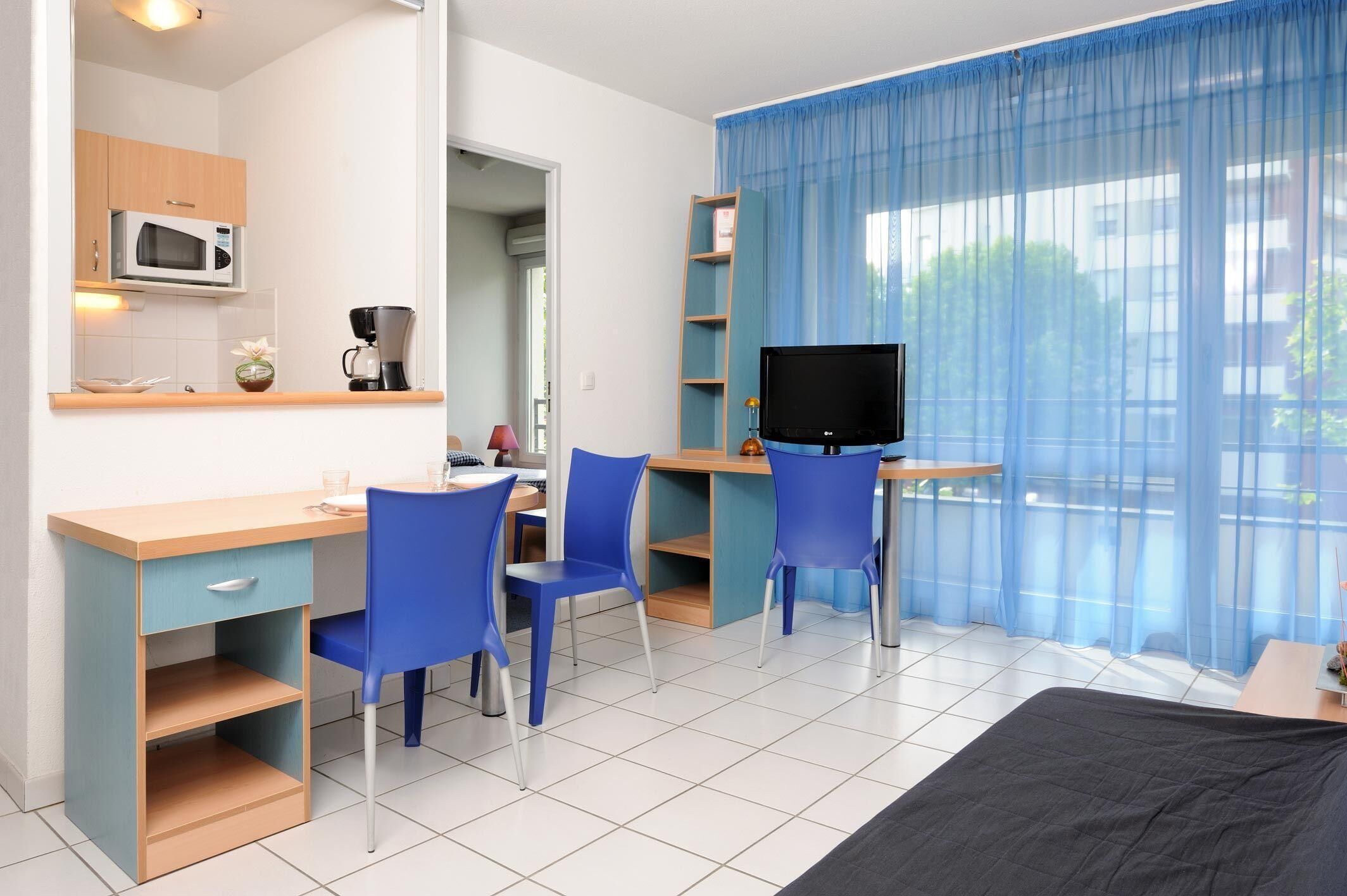 A compact hotel room with a bed, a desk with a chair, a television, and a kitchenette.