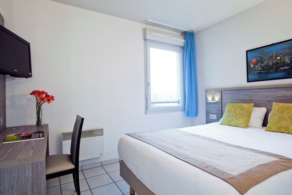 Ein Hotelzimmer mit Bett, Schreibtisch, Stuhl, Fernseher, einem Fenster mit Vorhängen und einem Bild an der Wand.