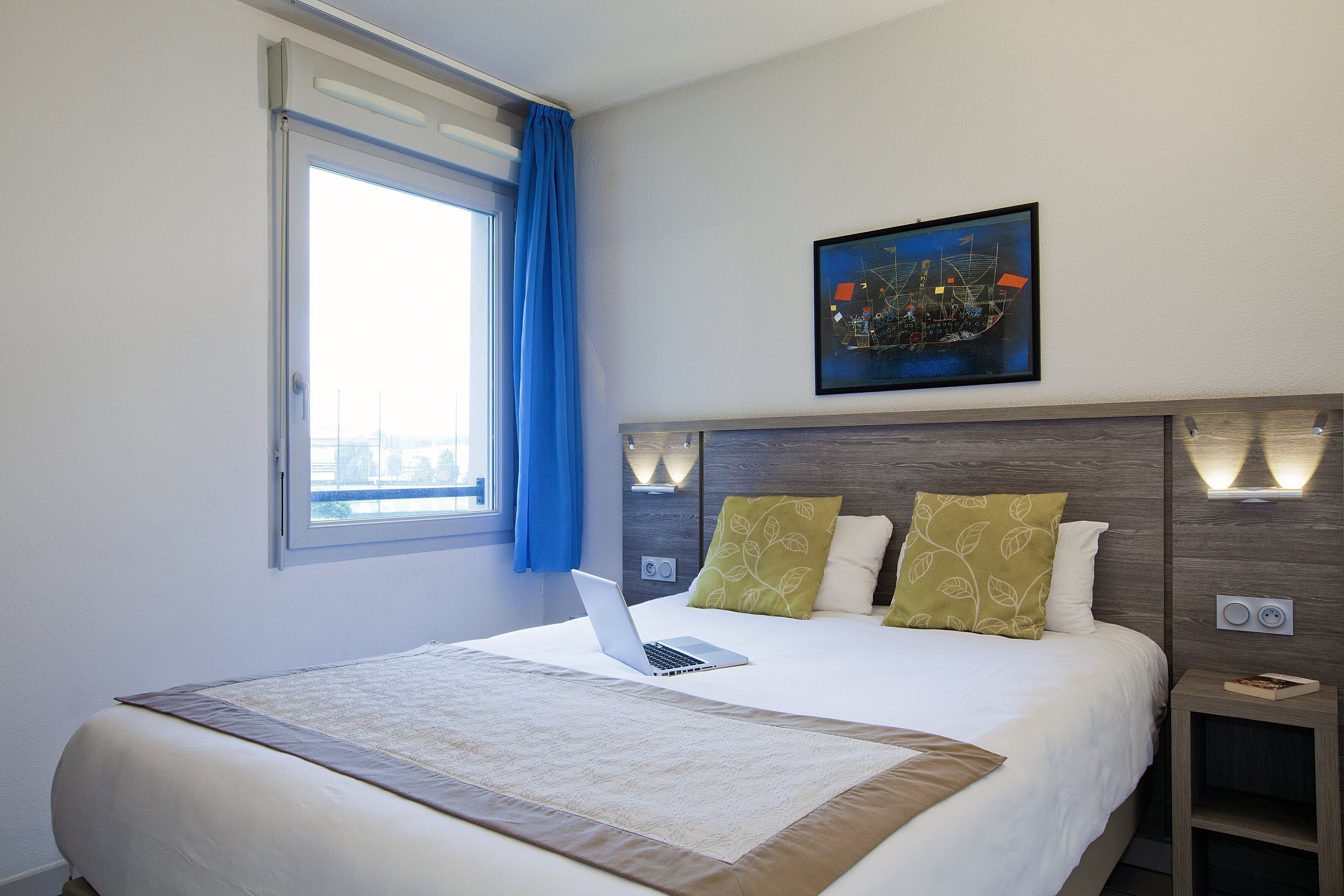 Una cama bien hecha con una computadora portátil, una ventana con cortinas azules y un cuadro en la pared.