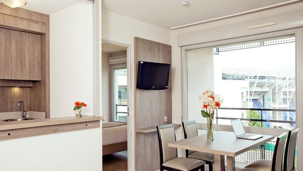 A modern room with a wooden kitchenette, a dining area with a table and chairs, a flat-screen TV mounted on the wall, and a balcony with a view of the city. - Séjours & Affaires Clermont-Ferrand Park Republique (Clermont-Ferrand)