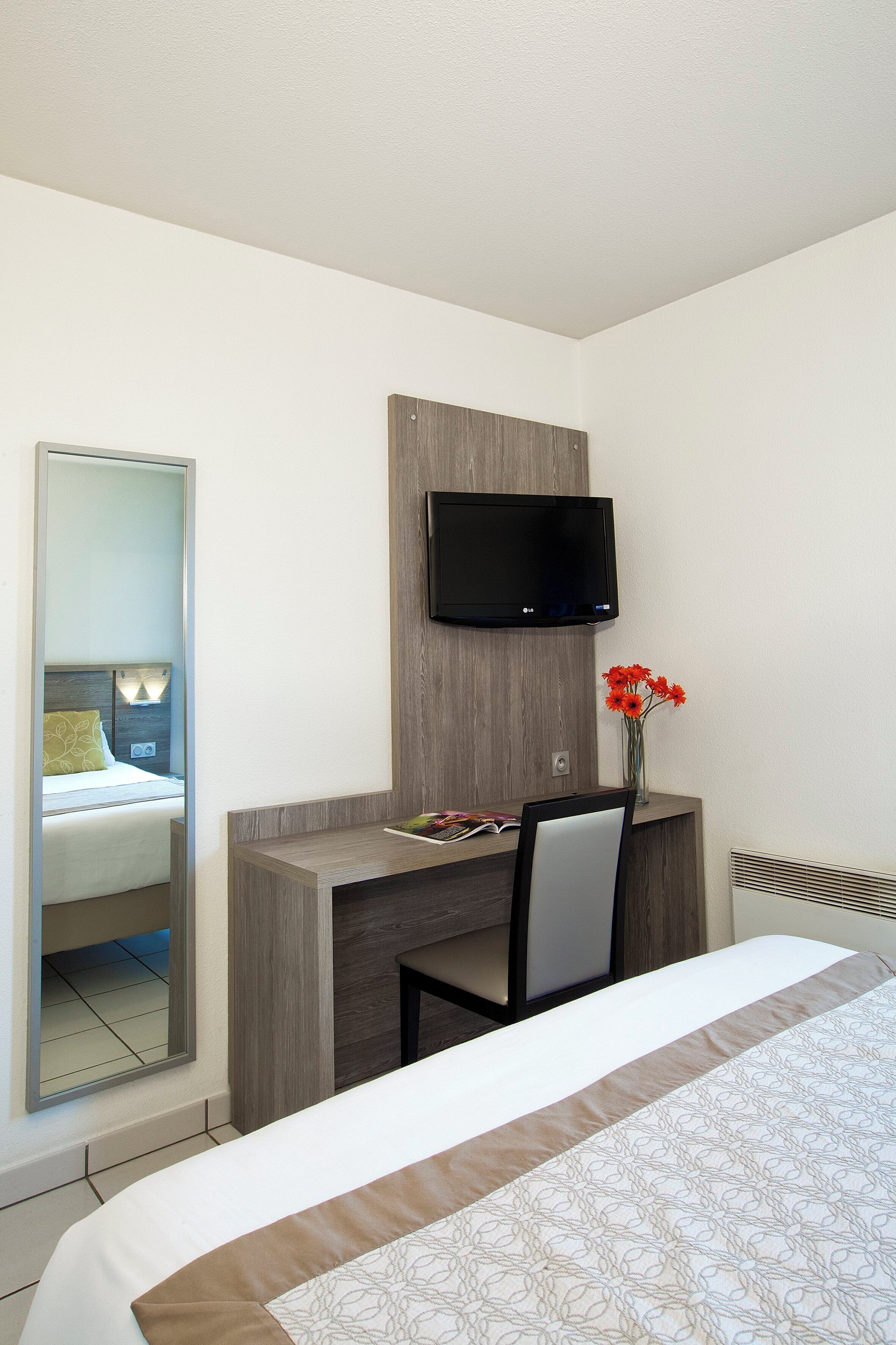 A hotel room with a bed, a desk with a chair, a TV mounted on the wall, and a mirror.