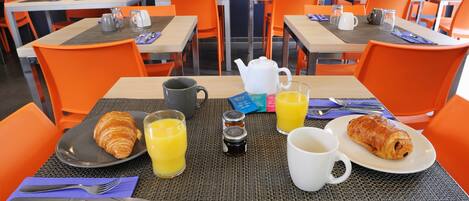 A breakfast setting with croissants, orange juice, and coffee in a dining area with orange chairs and tables.