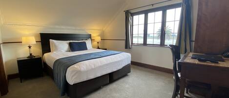 A hotel room with a large bed, two bedside tables with lamps, a desk with a chair, and a window with a view of palm trees.