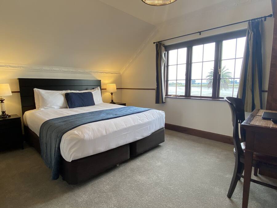 A hotel room with a large bed, two bedside tables with lamps, a desk with a chair, and a window with a view of palm trees.