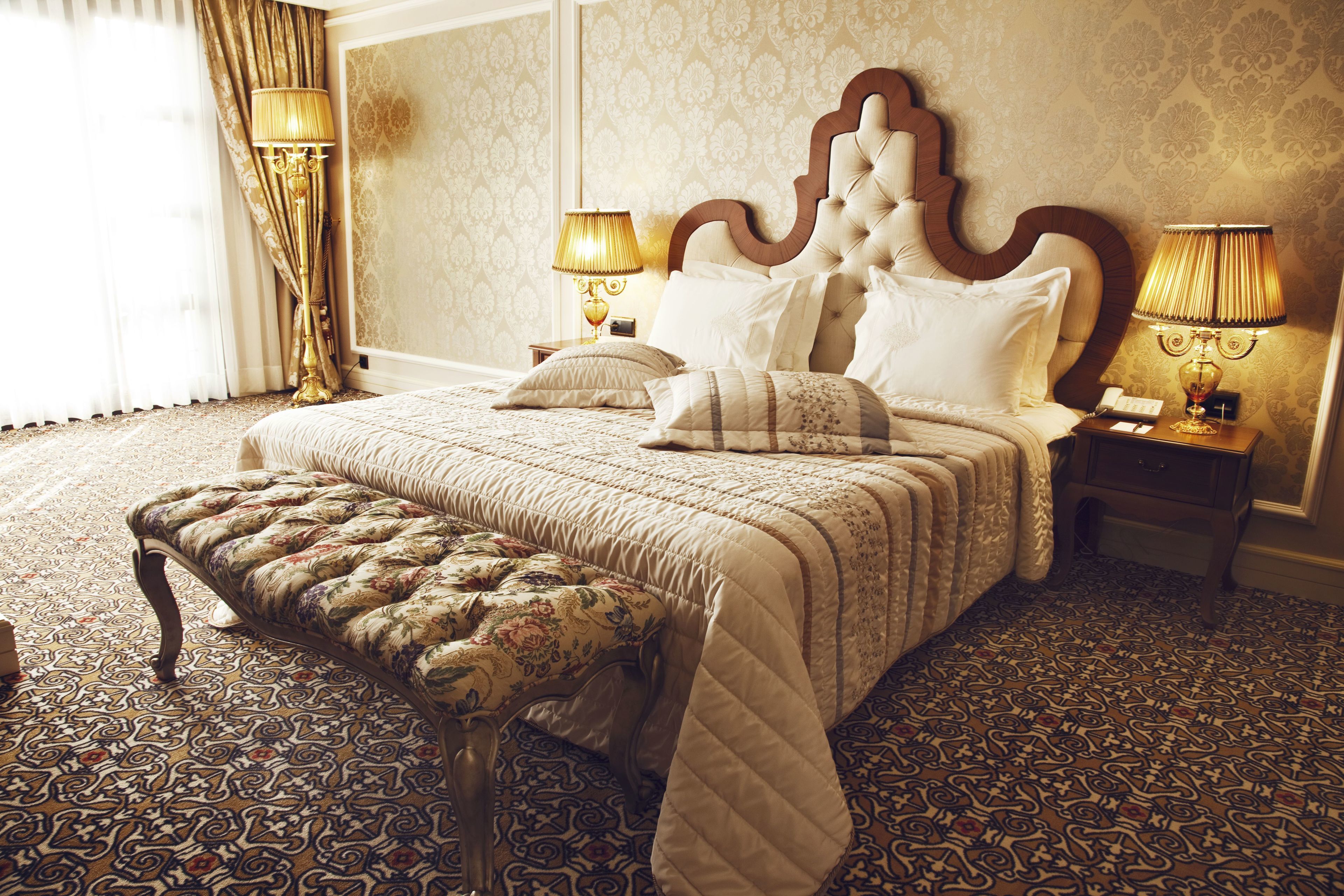 A large bed with a tufted headboard, two bedside tables with lamps, a bench, and patterned carpet.