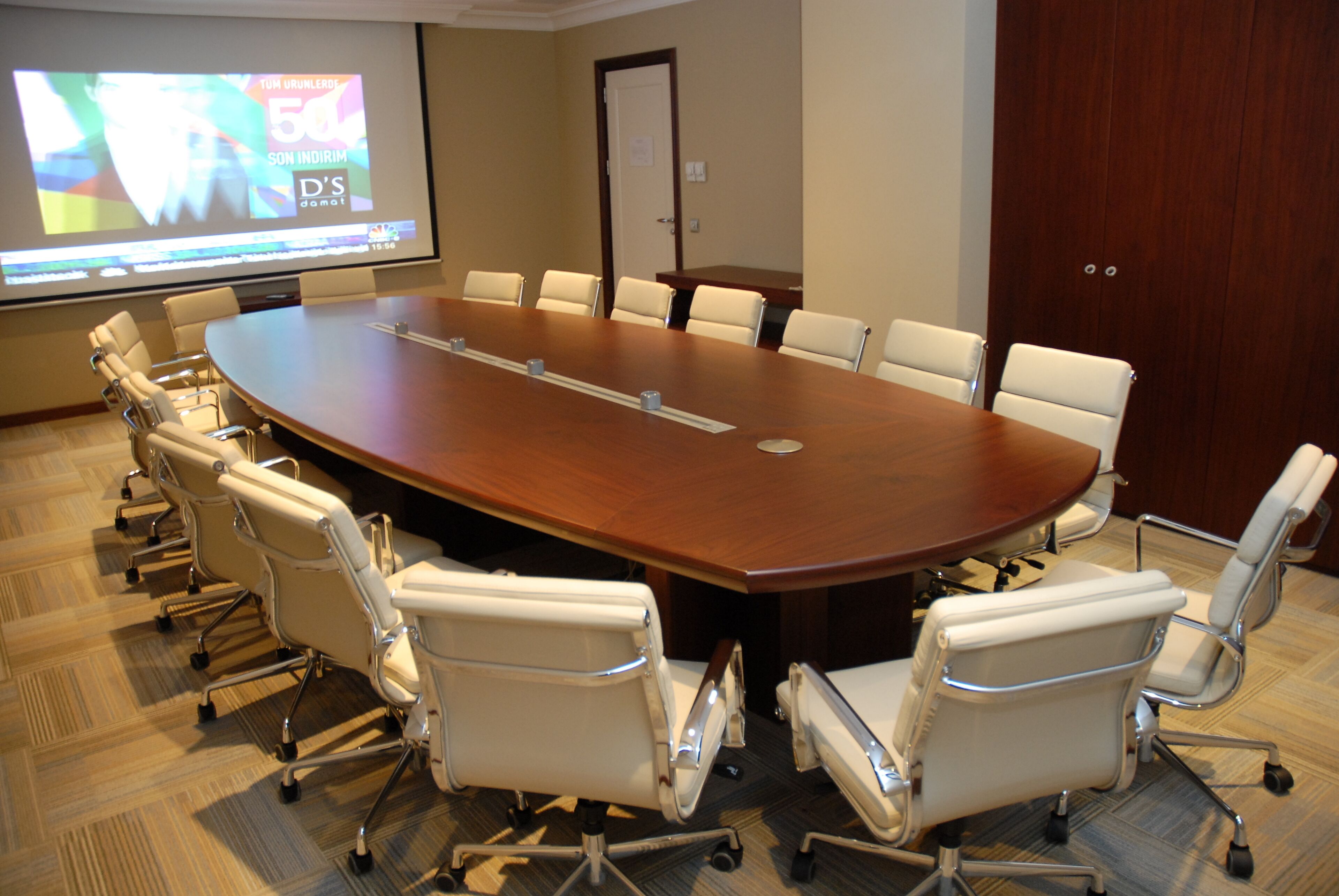 A conference room with a large oval table surrounded by white chairs, a projector screen, and a wooden door.