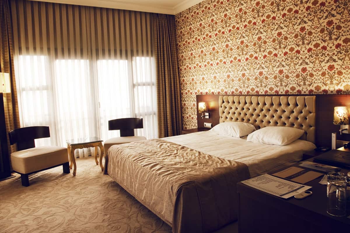 A hotel room with a large bed, a chair, a small table, and a nightstand with a lamp.