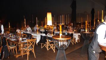 A restaurant with wooden tables and chairs, a central lamp, and a view of the water with tall structures.