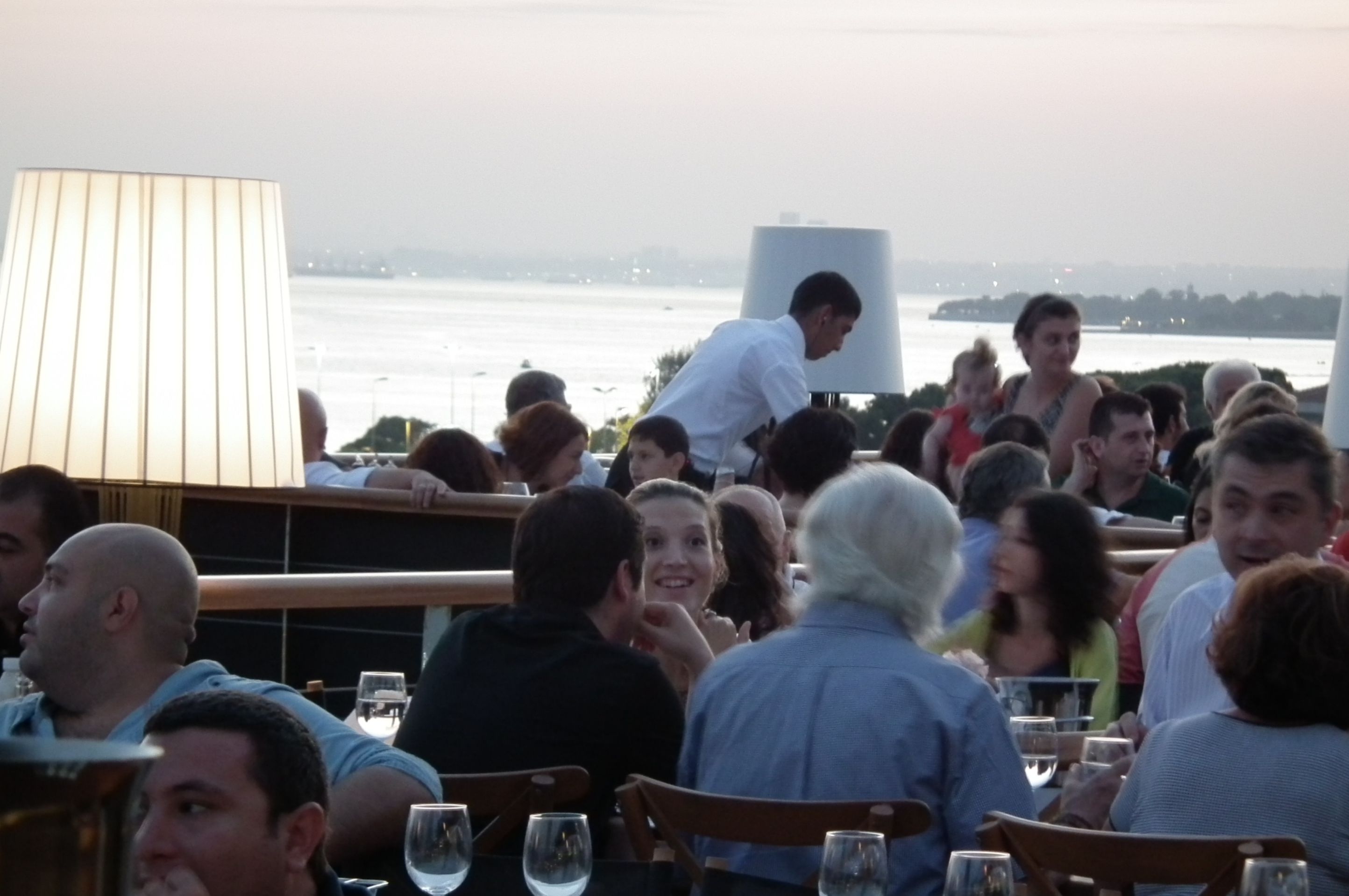 A crowded outdoor dining area with tables and chairs, overlooking a body of water.