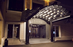 Entrada de hotel con puerta de vidrio, zona de acceso cubierta y escalera que lleva al piso superior.