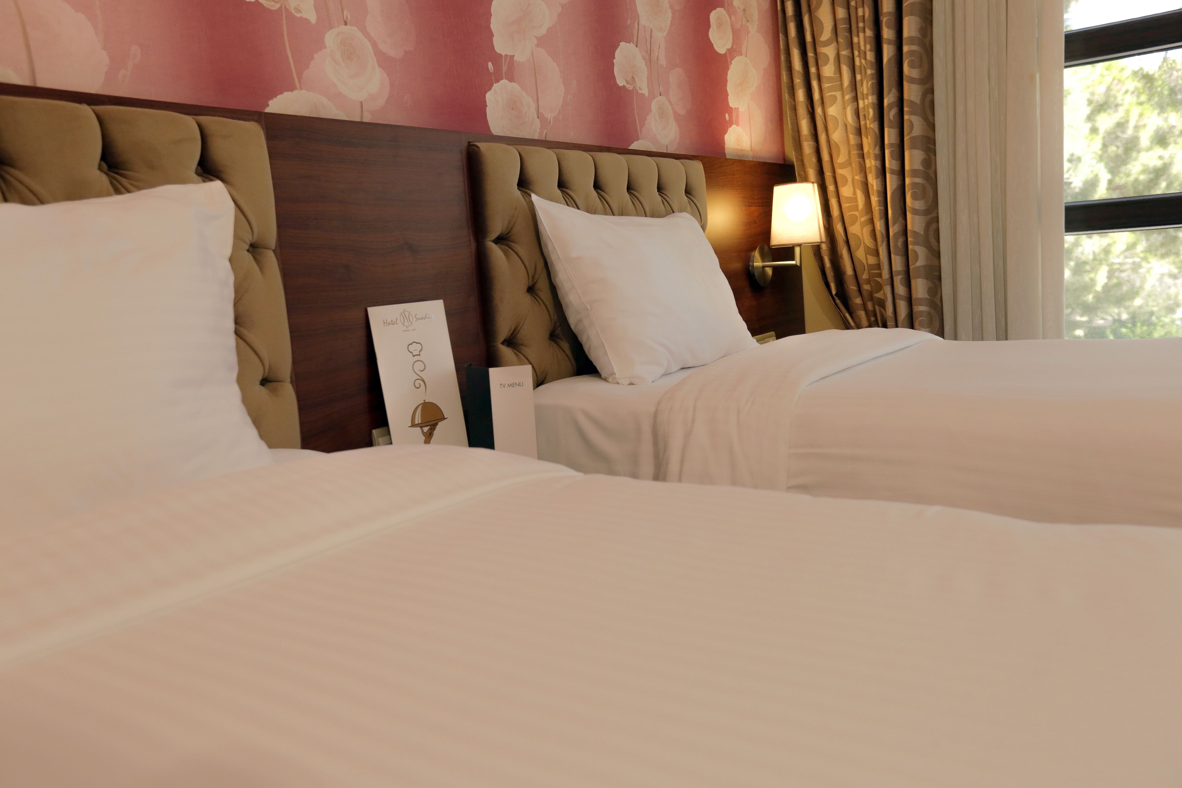 A hotel room with two beds, a wooden headboard, a nightstand with a lamp, and a wall with floral wallpaper.