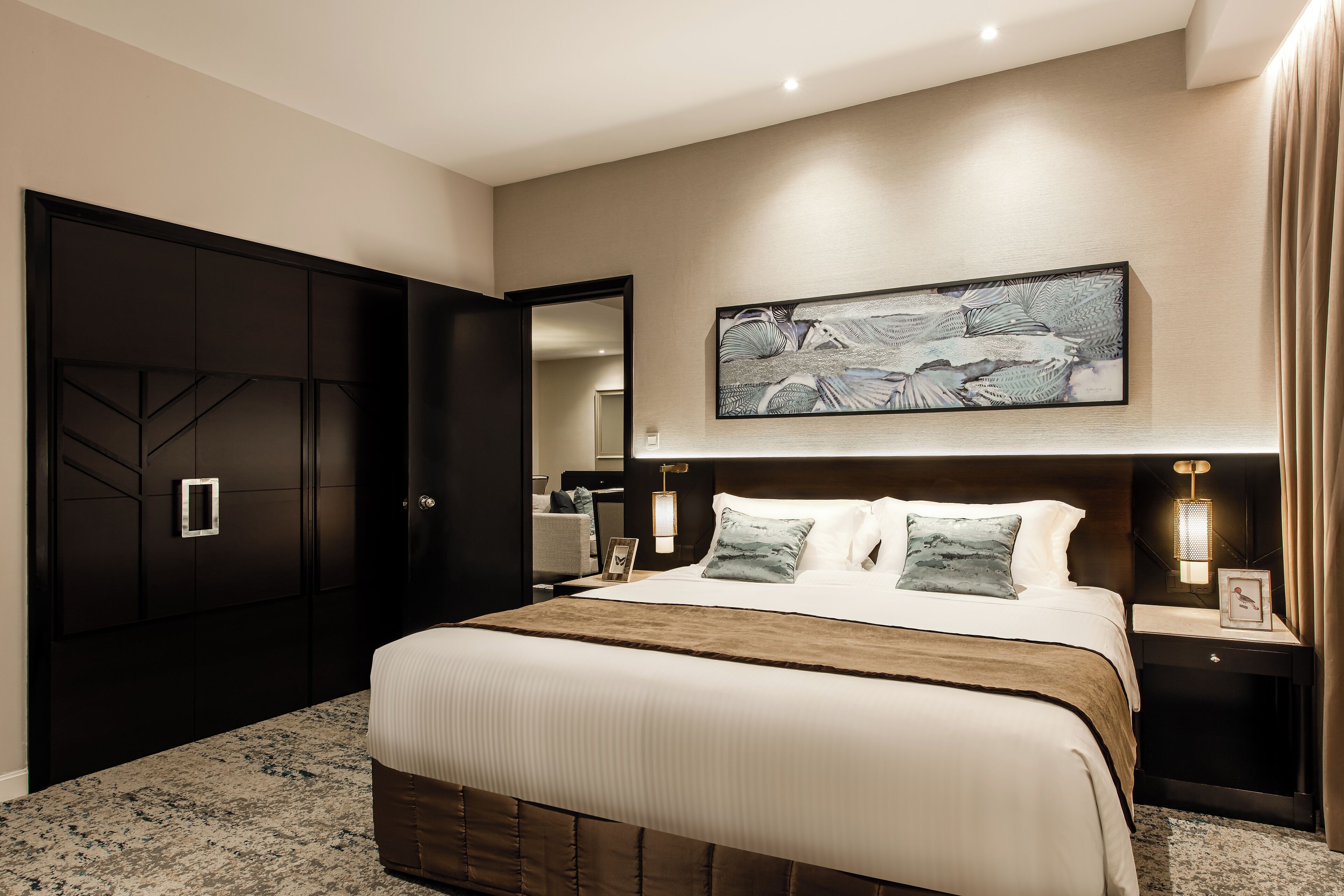 A modern hotel bedroom with a large bed, a dark wardrobe, a wall-mounted artwork, and a bedside table with a lamp.