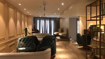 Un salón amplio con un sofá grande, dos sillones y una mesa de centro. El suelo es de madera, hay un ventanal con cortinas y una estantería decorativa con libros y una lámpara.