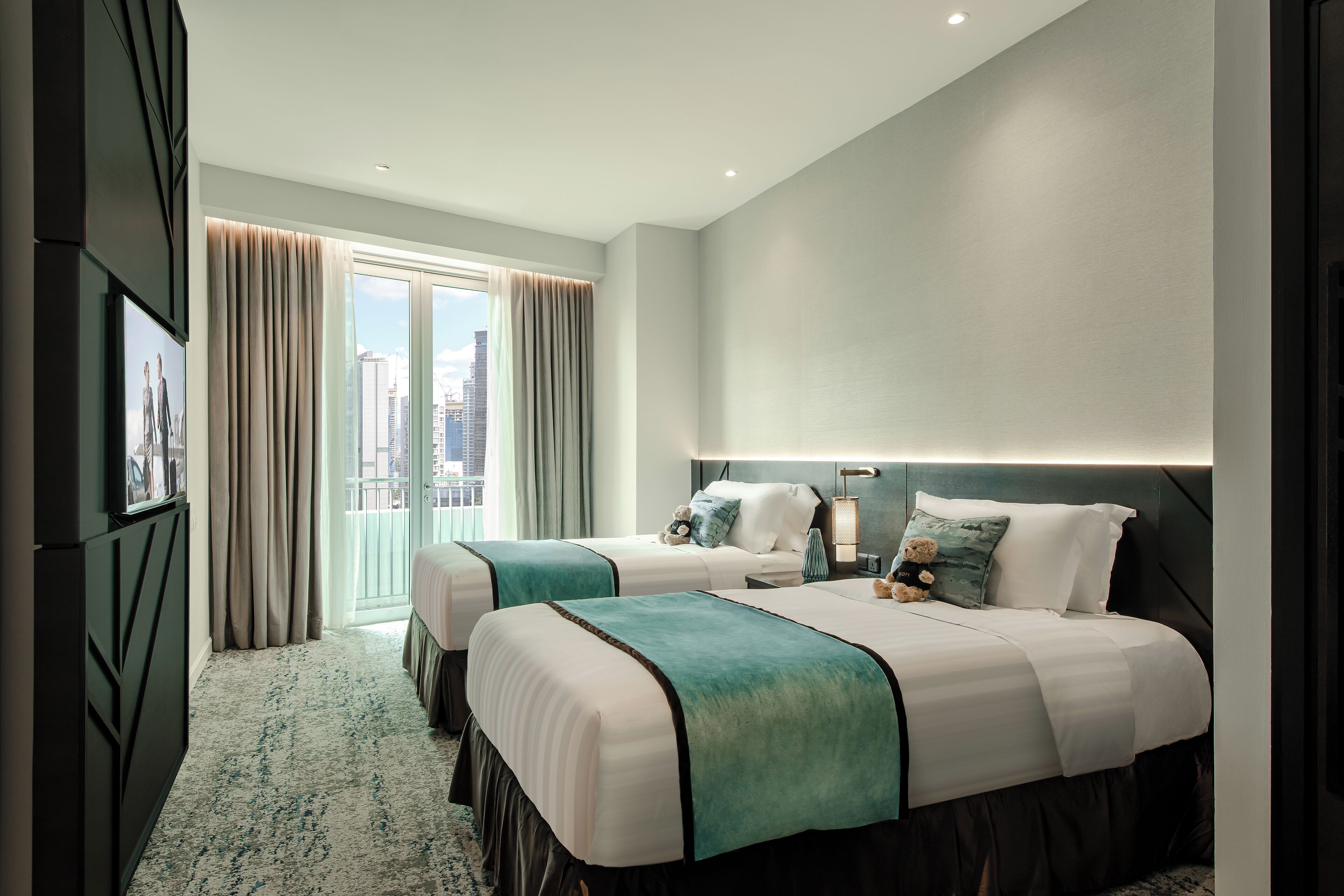 A modern hotel room with two beds, a large window with curtains, a city view, and a flat-screen TV mounted on the wall.